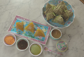 Image of rice dumplings laid out on colourful Peranakan tableware. There are three kee zhangs on the bottom left plate, and it is flanked by three different condiments that look like orange coconut sugar, gula melaka, and kaya.